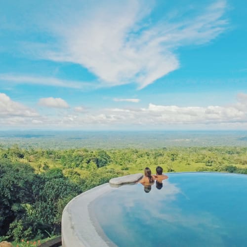 Zwei Personen entspannen sich in einem Infinity-Pool mit Blick auf üppiges Grün und einen teils bewölkten Himmel.