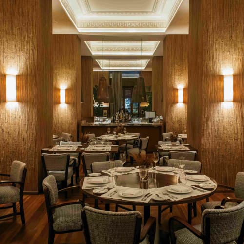 Elegant restaurant interior with round and rectangular tables set with white plates, glasses, and cutlery, surrounded by upholstered chairs, warm lighting, and wooden textured columns.