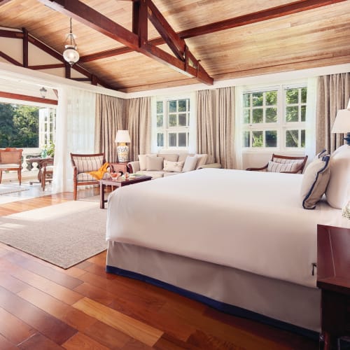 A spacious bedroom with a large bed, wooden ceiling, and windows overlooking a lush green outdoor area.