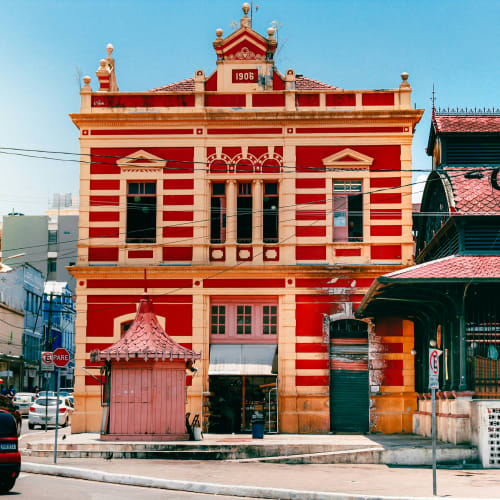 Bright red and beige historic building with '1906' displayed at the top, located on a street corner with cars nearby under a clear blue sky.