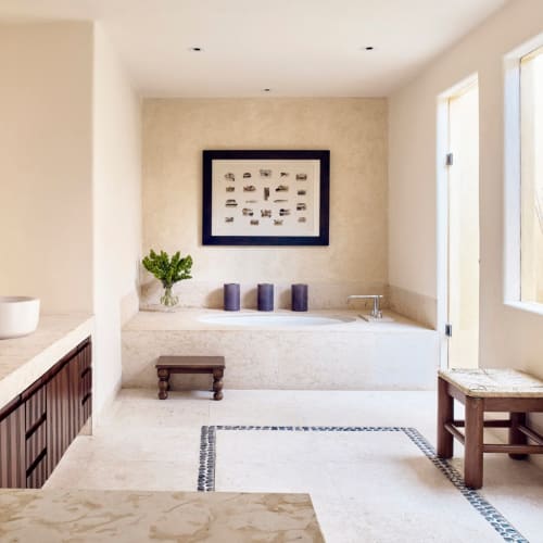 Großes, helles Badezimmer mit eingebauter Badewanne, zwei Aufsatzwaschbecken auf Marmorarbeitsplatte, Holzschemel, gerahmtem Kunstwerk und hellbeigen Wänden mit minimalistischer Dekoration.