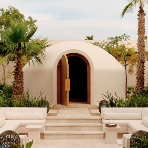 Ein gewölbtes, kuppelförmiges weißes Spa-Temazcal mit einer offenen Holztür, umgeben von Palmen und üppigem Grün, mit cremefarbenen Steinstufen und Außenbestuhlung davor.