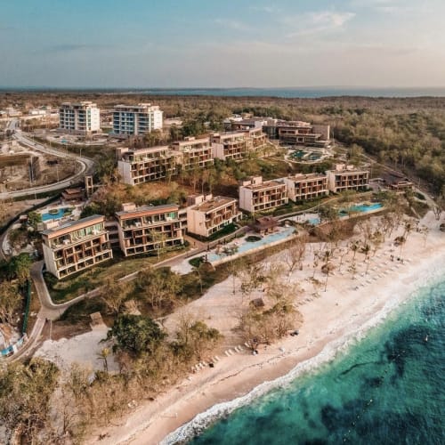 Luftaufnahme eines Strandresorts mit modernen Gebäuden, Schwimmbädern und Liegestühlen am Sandstrand mit türkisfarbenem Wasser unter leicht bewölktem Himmel.