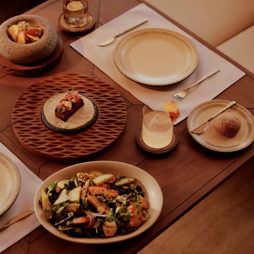 Ein Holztisch mit verschiedenen Gerichten, darunter ein Salat in einer Schüssel, ein angerichtetes Dessert, eine Schüssel mit Obstscheiben, Brot auf einem kleinen Teller und zwei Gläser mit Getränken, eines mit getrockneter Zitrusscheibe garniert.
