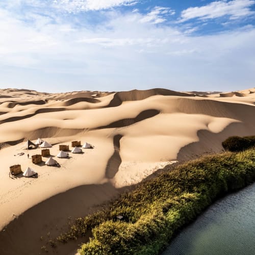Ein kleines Wüstencamp mit weißen Zelten zwischen Sanddünen neben einem grünen, bewachsenen Flussufer unter einem teilweise bewölkten blauen Himmel.