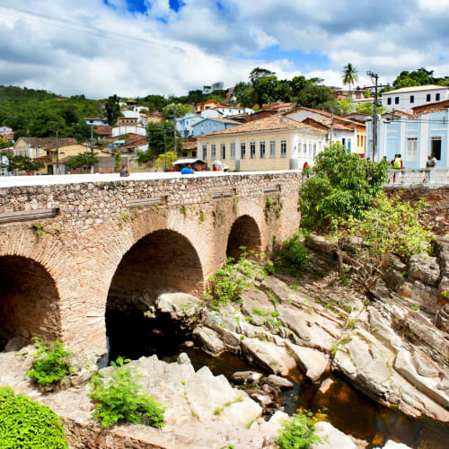 Eine historische Steinbrücke spannt sich über ein felsiges Flussbett in einer bunten Kleinstadt mit traditionellen Häusern am Hügel.