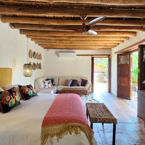 Spacious rustic bedroom with wooden ceiling beams, a large bed with colorful cushions and a pink throw, a wooden dresser with a flat screen TV, and an open door leading to an outdoor area.