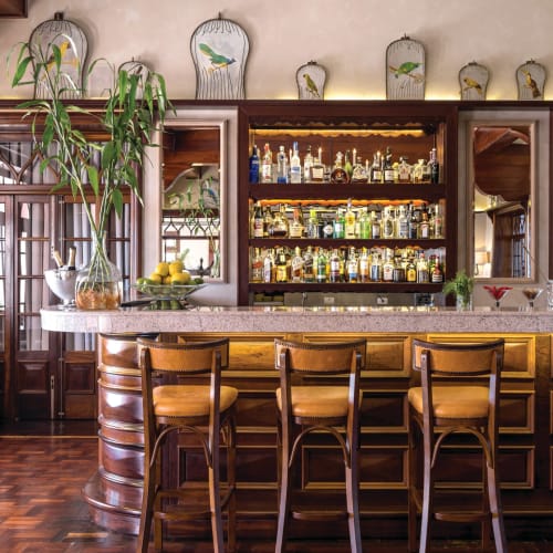 Eine luxuriöse Bar mit drei hölzernen Stühlen davor, einer Marmor-Arbeitsplatte und einem gut bestückten Spirituosenregal dahinter, dekoriert mit Topfpflanzen, Zitronen und vogelähnlichen Ornamente auf dem Regal oberhalb.