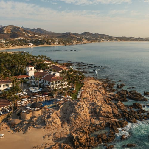 Luftaufnahme des Resorts One&Only Palmilla am Morgen mit Gebäuden, Schwimmbad, Palmen, felsiger Küste und ruhigem Meer unter leicht bewölktem Himmel.