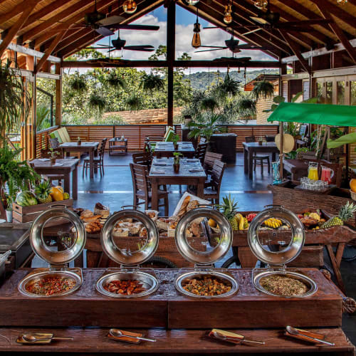 Ein geräumiges Restaurant in Arenal, Costa Rica, für das Frühstück vorbereitet mit einer Auswahl an Speisen auf einem Buffet, umgeben von Pflanzen und Holzakzenten.