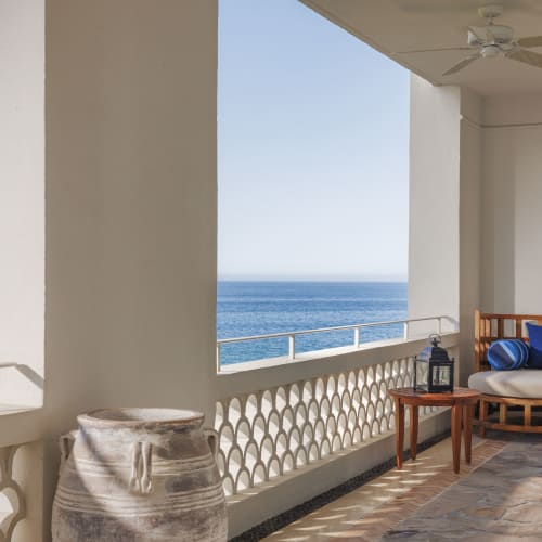 Ozeanfront-Terrasse einer Luxussuite mit weißem Stuckgeländer, einem gepolsterten Holzsofa mit blauen und weißen Kissen, einem kleinen Holztisch mit Laterne und einer großen Keramikvase mit Blick auf das ruhige Meer unter klarem Himmel.