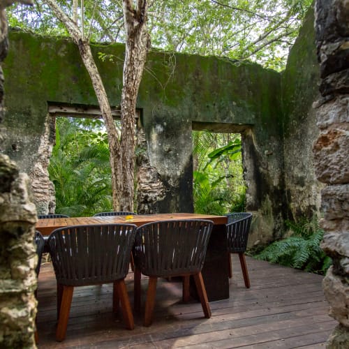 Holztisch mit sechs Stühlen auf einem Holzdeck in einer von Moos bedeckten Steinhütte, umgeben von üppigem Grün im Yucatan.