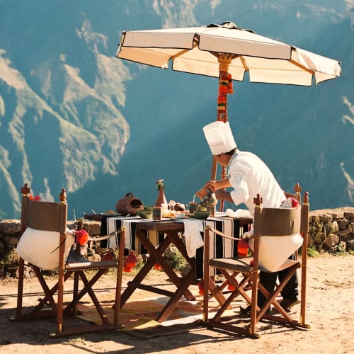 Ein Koch mit weißem Hut und Jacke bereitet Essen an einem Esstisch im Freien unter einem großen Sonnenschirm vor, im Hintergrund Berge.