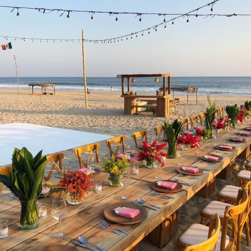 Ein langer Holztisch, der für ein Abendessen am Strand mit Sand und Ozeanwellen im Hintergrund gedeckt ist. Der Tisch ist mit Blumen, Glaswaren und Tellern dekoriert, und darüber hängen Lichterketten.