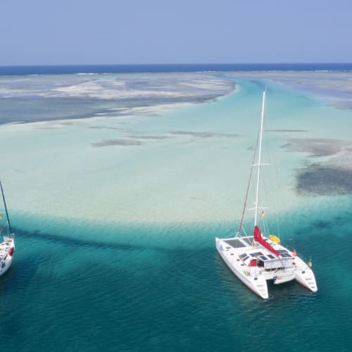 Two sailboats are anchored in the turquoise waters near a white sandy island in the Caribbean.