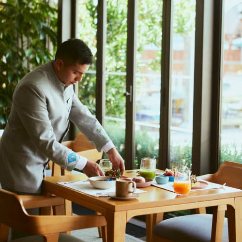 A chef arranging food on a table in a bright, modern restaurant with large windows and greenery outside.