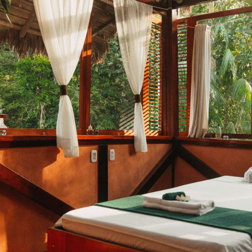 Massageraum in einer Holzhütte mit großen Fenstern, die üppiges grünes Laub draußen zeigen, ausgestattet mit einer Massageliege mit gefalteten Handtüchern und weißen Vorhängen.