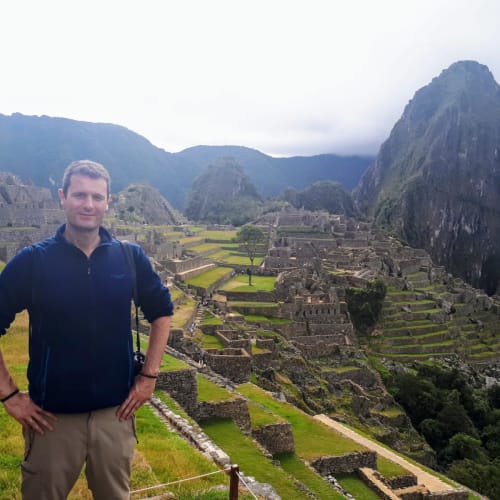 Mann steht mit den Händen in den Hüften vor den alten Stein-Terrassen und Ruinen von Machu Picchu, mit Bergen im Hintergrund unter bewölktem Himmel.