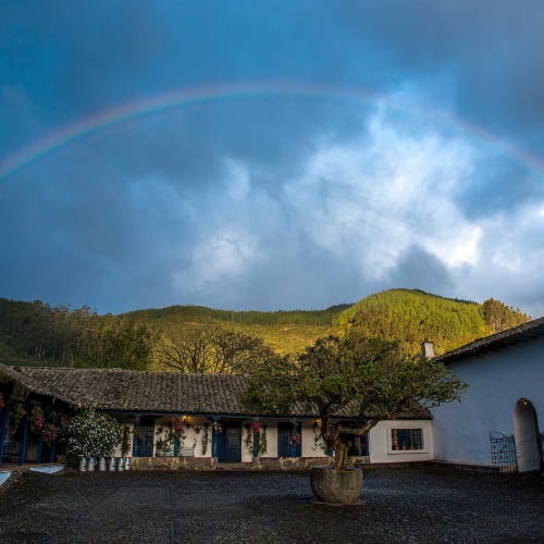 Ein Innenhof einer traditionellen Hacienda mit einem zentralen Baum in einem Topf, weißen Gebäuden mit Ziegeldächern, einer bergigen Landschaft im Hintergrund und einem Regenbogen, der sich über den wolkigen Himmel spannt.