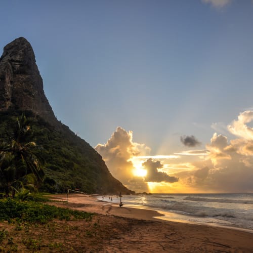 Ein malerischer Strand bei Sonnenuntergang mit einem hohen felsigen Hügel auf der linken Seite und dem Ozean auf der rechten Seite, Sonnenstrahlen durchbrechen Wolken.