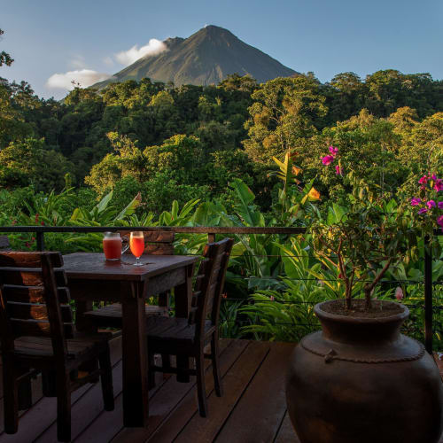 Terrasse eines Restaurants mit einem Holztisch und Stühlen, mit Blick auf üppiges grünes Laub und einen Vulkan im Hintergrund.