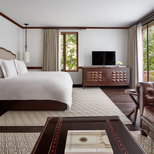 Elegante Suite in einem Four Seasons Hotel mit großem Bett, gemusterten Teppichen, einem Holzschrank mit Fernseher und zwei Fenstern mit Blick auf Grünflächen.