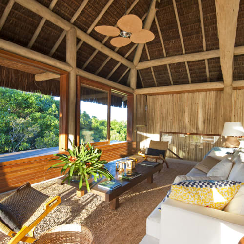 Eine gemütliche Lounge mit großen Fenstern mit Blick auf üppig grüne Bäume, eingerichtet mit einem weißen Sofa, Sesseln, Pflanzen und einem Deckenventilator.