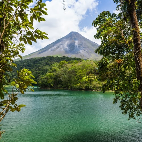 A serene lake surrounded by lush green trees with a volcano in the background under a partly cloudy sky.