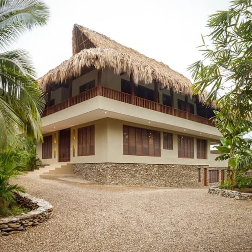 Two-story villa with a thatched roof and stone base surrounded by tropical plants and palms, under a bright sky.
