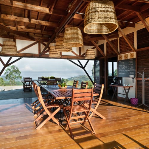 A rustic open wooden dining area with a table and chairs set, featuring hanging woven lamps and a view of trees and hills through large open windows.