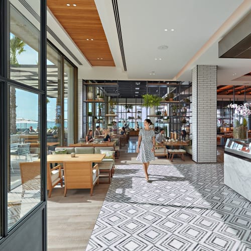 Ein modernes, gehobenes Restaurant mit großen Glasfenstern, die auf eine Palme draußen blicken, mit einem Barista, der Getränke an einer Marmortheke zubereitet.