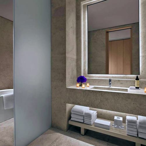 A modern bathroom with a bathtub on the left, a large illuminated mirror above a sink, and neatly folded towels on a shelf below.