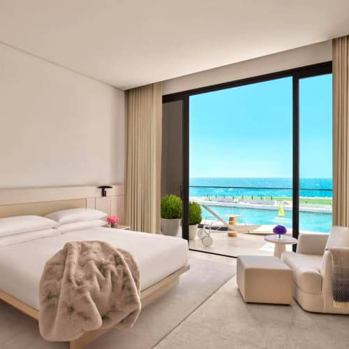 A luxurious hotel room with a balcony overlooking the sea, featuring a large bed with plush blankets, a sitting area with a chair and ottoman, and elegant decor.