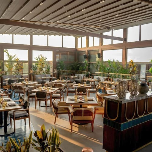 Elegantes Innenrestaurant mit großen Fenstern, gedeckte Tische mit Gläsern und Besteck, dekoriert mit Topfpflanzen unter einer Holzboden-Decke.