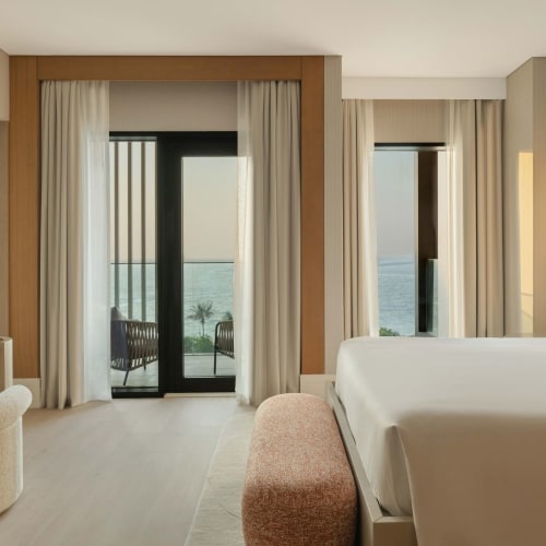 An elegant hotel room with a balcony overlooking the sea, featuring a large bed, a sitting area, and modern decor.