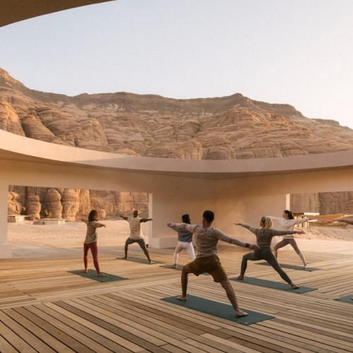 Eine Gruppe von Menschen, die Yoga in einem modernen Gebäude mit großen kreisförmigen Öffnungen mit Blick auf eine Canyon-Landschaft draußen praktiziert.