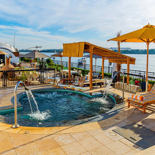 Outdoor pool area with water fountains, wooden pergola bar, striped lounge chairs, and yellow umbrellas overlooking a river under a partly cloudy sky.
