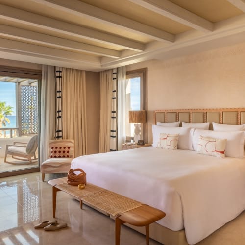 Luxurious seaside hotel room with a large bed, nightstands, and a balcony view of the beach and palm trees.