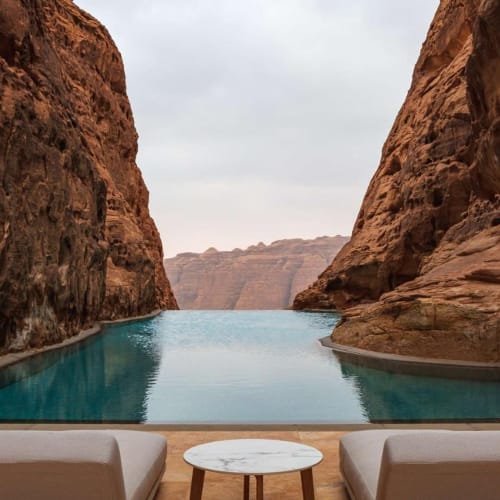 A luxurious pool with a view of rocky canyon cliffs, two lounge chairs, and a small round table in front of it.