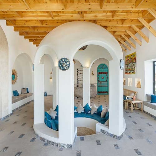 Interior of a lounge with white arched walls, blue cushions in a circular sunken seating area, wooden ceiling beams, decorative plates, and a window showing outdoor landscape.