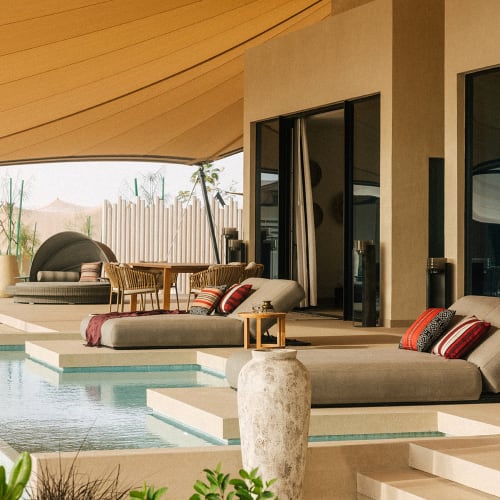 Luxury villa with a pool, outdoor lounge furniture, and a large shaded area with a wooden roof, overlooking a garden with green plants.