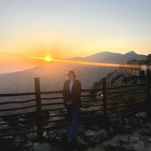 Person standing by a wooden fence on a rocky path watching the orange sun setting behind mountain peaks.