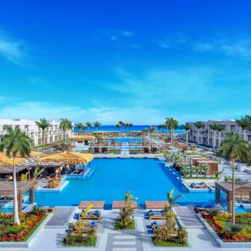 Ein luxuriöses Resort mit einem großen Swimmingpool, umgeben von Palmen, Blumenbeeten und weißen Gebäuden, mit Blick auf das Meer unter einem klaren blauen Himmel.