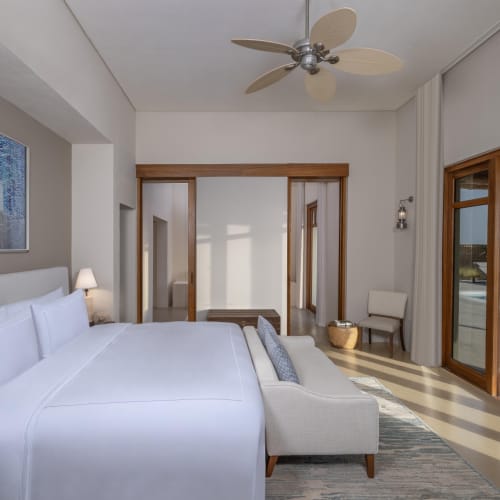 Helles, modernes Resort-Schlafzimmer mit großem weißen Bett, Deckenventilator und Glastüren, die zu einem Außenpool mit schönem Ausblick führen.