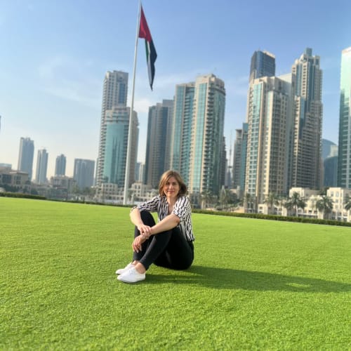 Eine Frau sitzt auf einer grünen Rasenfläche mit der Skyline von Dubai und einer hohen Fahnenstange mit der Flagge der VAE im Hintergrund unter klarem blauem Himmel.