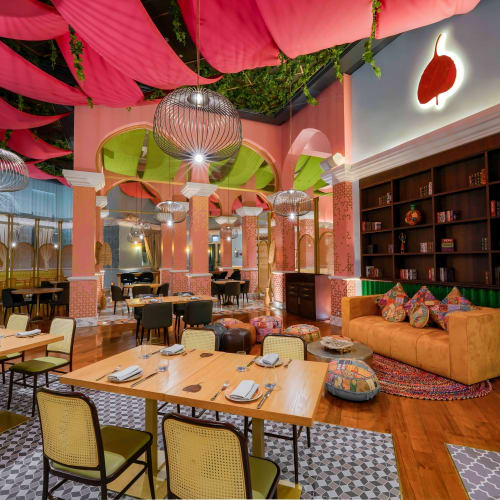 Ein stilvolles Restaurantinterieur mit rosa drapierter Decke, grünen und rosa Säulen, Holztischen mit gedeckter Tafel, gelben und schwarzen Stühlen, einem senfgelben Sofa mit gemusterten Kissen und einem großen Bücherregal mit Dekorationsgegenständen.