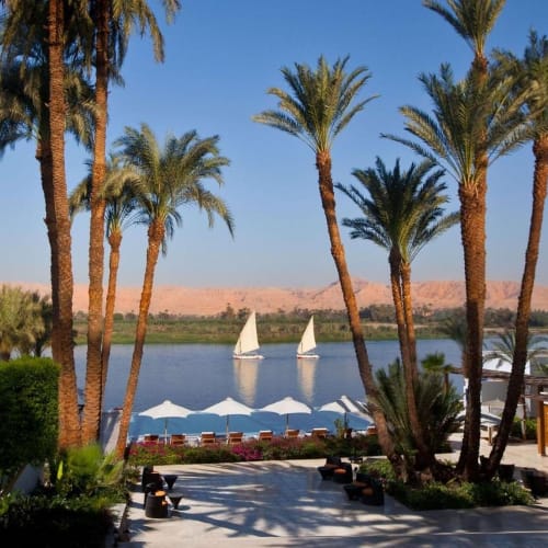 A view of the Nile River at Hilton Luxor Resort & Spa with sailboats, palm trees, and a sandy landscape in the background.