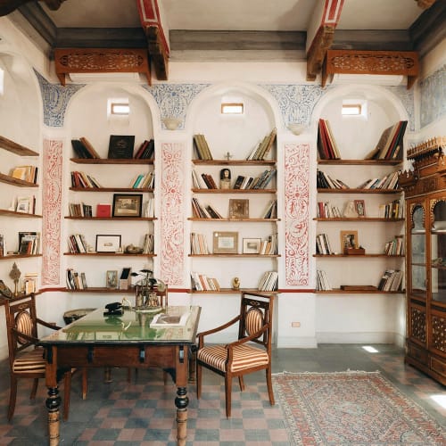 Ein gemütliches Bibliothekszimmer mit weißen Regalen voller Bücher, einem Holztisch mit Stühlen und einem gemusterten Teppich auf einem Fliesenboden.