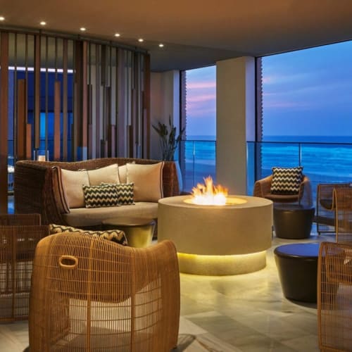Luxurious hotel lounge with a central fireplace, wicker chairs, and a view of the ocean at sunset.