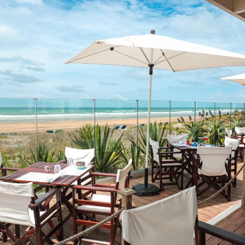 Outdoor dining area at a beachside resort with wooden tables, white chairs, large white umbrellas, and a glass fence overlooking the sandy beach and ocean under a partly cloudy sky.